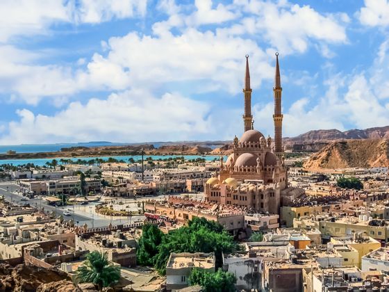 Vista elevada de una ciudad con una gran mezquita de dos minaretes, con agua azul y colinas áridas en el fondo bajo un cielo nublado.