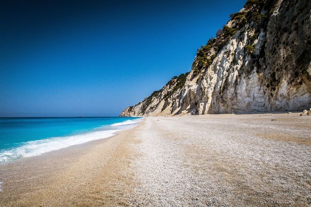 Un'ampia spiaggia di ciottoli incontra acque turchesi accanto a una scogliera rocciosa a picco sotto un cielo azzurro e limpido.
