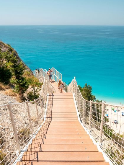 Due persone scendono una lunga scalinata in legno costruita nella scogliera, che porta a una spiaggia sabbiosa con acqua turchese.
