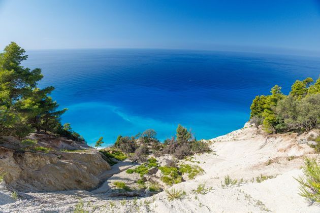 Una vista grandangolare da una scogliera sabbiosa e alberata che si affaccia sul mare blu e turchese vivido sotto un cielo sereno.