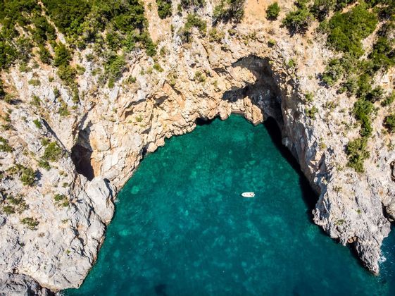 Una vista aerea di una piccola barca bianca che galleggia in una cala turchese circondata da scogliere rocciose e alberi verdi.