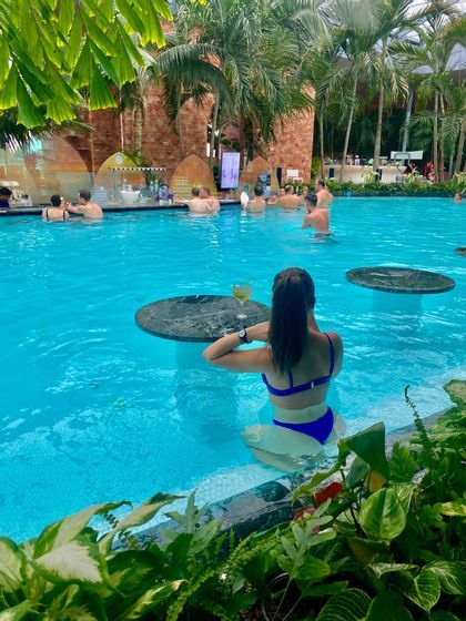 Una mujer en bikini azul se sienta en una mesa en una piscina con un bar acuático rodeada de palmeras.