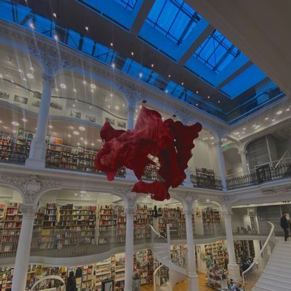 Una gran instalación de arte rojo cuelga del techo de una librería de varios niveles con columnas blancas ornamentadas y un tragaluz de cristal.