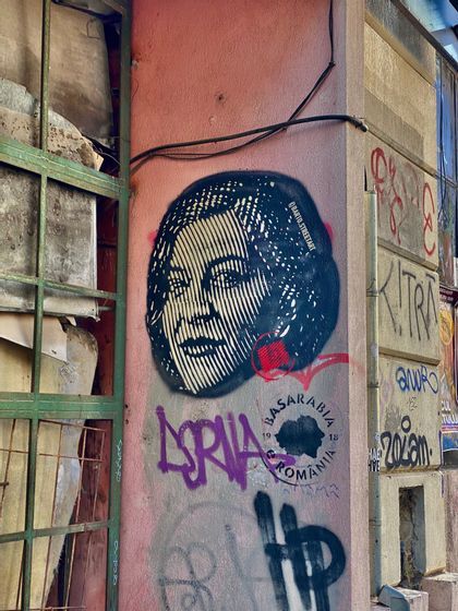 Un esténcil del rostro de una mujer pintado con aerosol en una pared urbana rosa, cubierta con varios grafitis coloridos.
