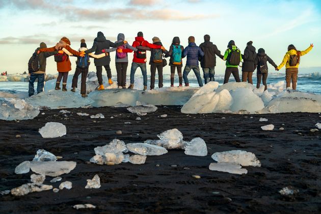 Un grupo de WeRoad con los brazos entrelazados sobre grandes bloques de hielo en una playa de arena negra, frente al océano.