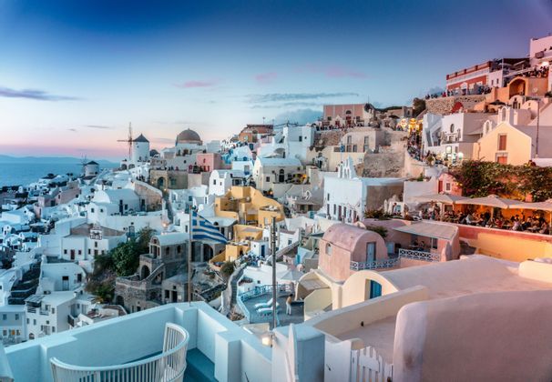 Un villaggio greco con edifici bianchi e mulini a vento arroccato su una scogliera a picco sull'oceano al tramonto.