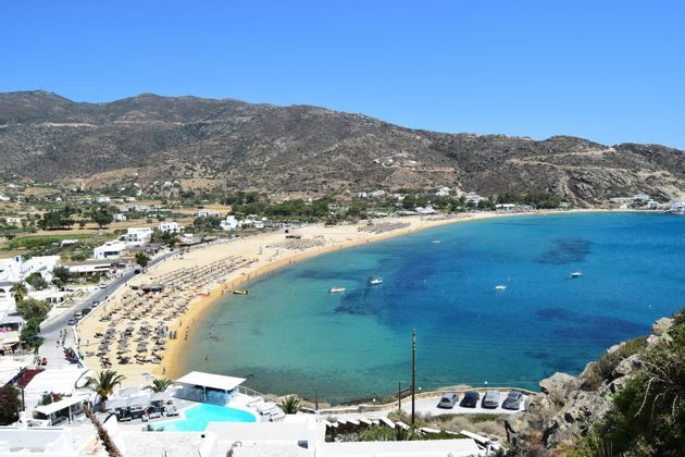 Una vista dall'alto di una spiaggia sabbiosa affollata che curva attorno a una baia calma e turchese, con colline che si ergono sullo sfondo sotto un cielo azzurro chiaro.