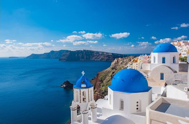 Edifici bianchi con cupole blu vivace si affacciano sul mare da una scogliera sotto un cielo sereno con nuvole sparse.