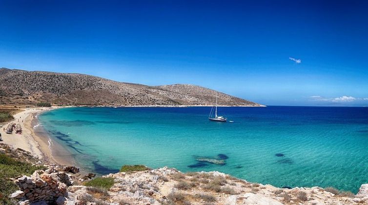 Una barca a vela ancorata in una baia con acqua limpida e turchese, una spiaggia di sabbia e colline rocciose sotto un cielo azzurro brillante.