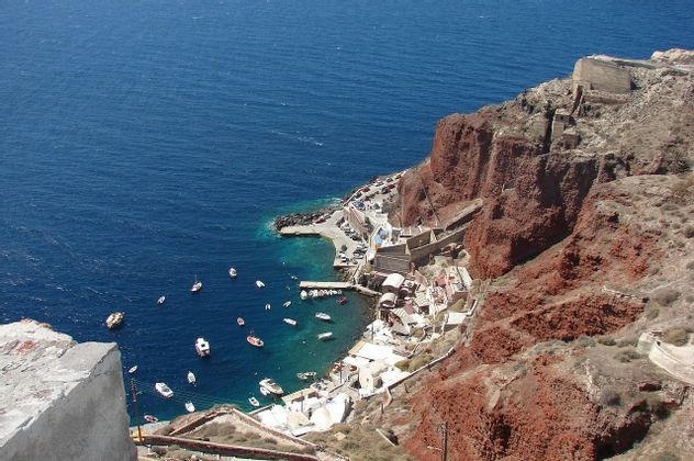 Una veduta dall'alto di un piccolo porto con barche ed edifici bianchi, adagiato ai piedi di una scogliera rossa e affacciato sul mare blu intenso.