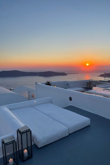 Lettini bianchi su una terrazza con vista sul mare e isole lontane mentre il sole tramonta all'orizzonte.