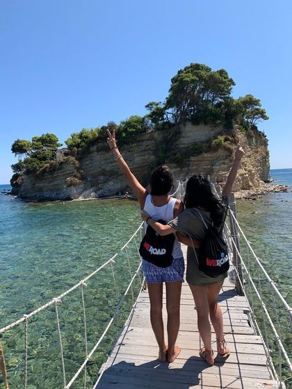 Deux femmes d'un voyage en groupe WeRoad se tiennent sur un pont de singe au-dessus d'une eau claire, faisant des signes de paix vers une petite île.