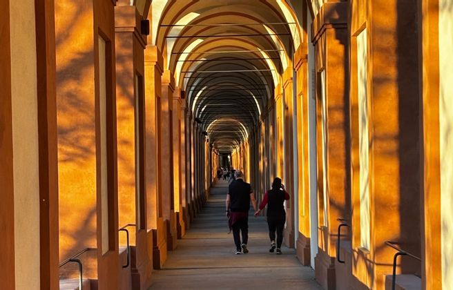 Una pareja camina de la mano por un largo pórtico arqueado mientras la cálida luz del sol proyecta largas sombras sobre las columnas.