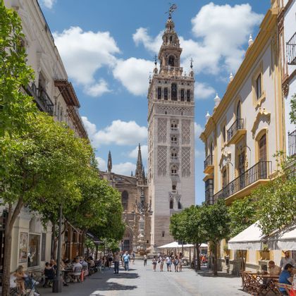 Un campanario ornamentado se alza al final de una soleada calle europea bordeada de árboles, cafés y gente paseando bajo un cielo azul.