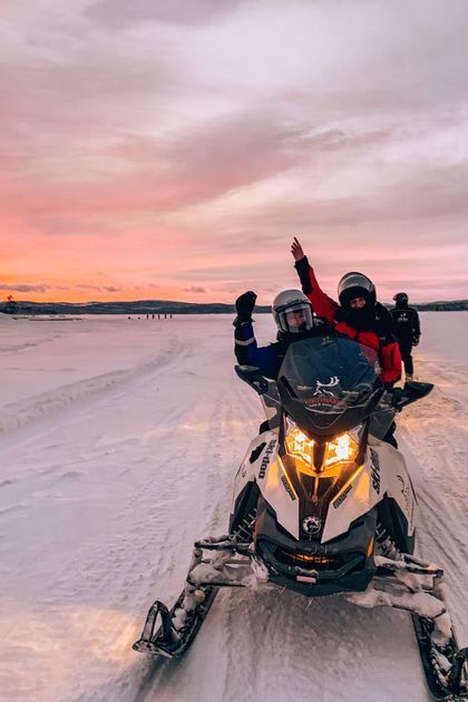 Two people from a WeRoad group trip wave from a snowmobile in a vast, snowy landscape under a pink sunset sky.