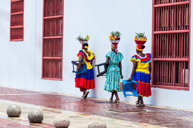 Tres mujeres con vestidos coloridos caminan por una acera empedrada, balanceando cuencos de fruta sobre sus cabezas y llevando taburetes pequeños.