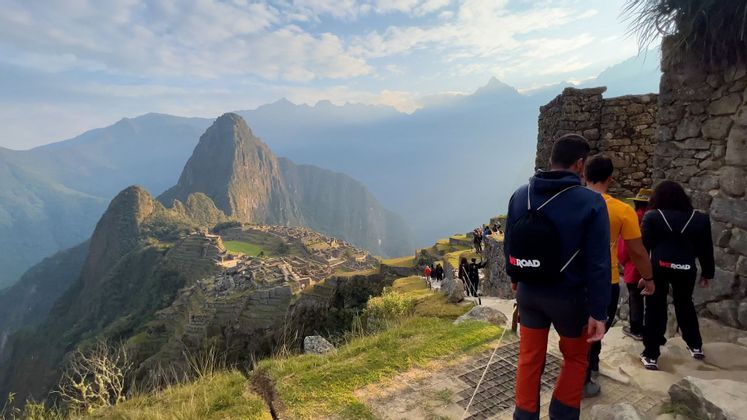 Un viaggio di gruppo WeRoad percorre un sentiero di pietra che si affaccia su antiche rovine, su un fianco di montagna, sotto un cielo parzialmente nuvoloso.