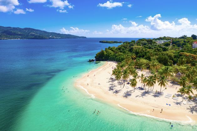 Una vista aérea de una playa tropical con gente nadando en agua turquesa y relajándose en arena blanca junto a exuberantes palmeras.