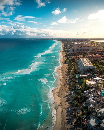 Vista aérea de una playa de arena atestada con agua turquesa, flanqueada por hoteles y palmeras bajo un cielo azul con nubes blancas.