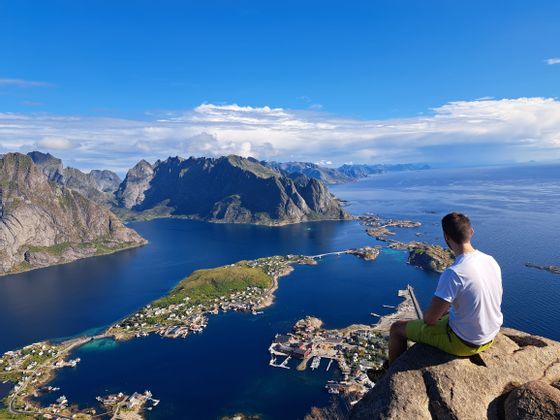 Un uomo siede su una roccia che domina un villaggio costiero, montagne e un fiordo sotto un cielo blu.
