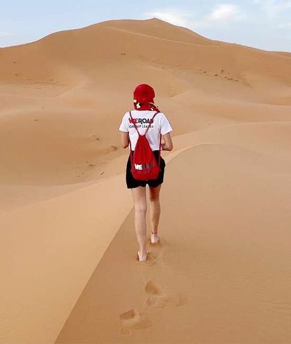A WeRoad group leader, seen from behind, walks barefoot on the ridge of a large sand dune, leaving footprints in the desert.
