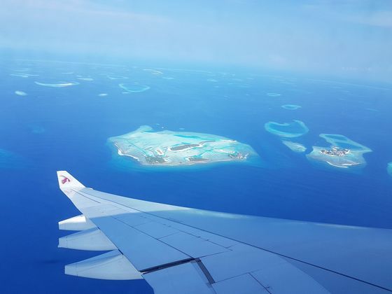 An aerial view from an airplane window showing the wing over a deep blue ocean scattered with small tropical islands and atolls.