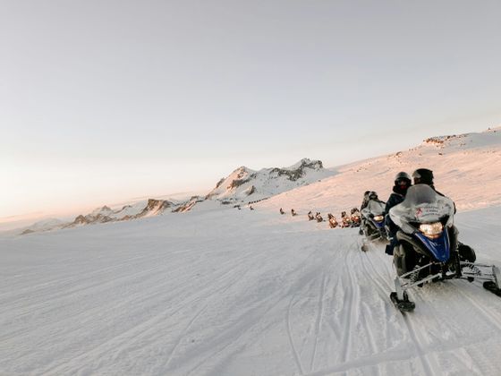 Un viaggio di gruppo WeRoad in motoslitta si snoda in fila attraverso un vasto paesaggio montano innevato al tramonto.