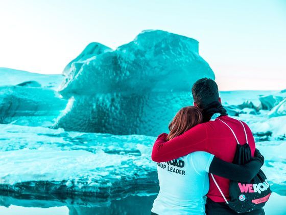 Two members of a WeRoad group trip hug while looking at a large blue iceberg in a frozen landscape.