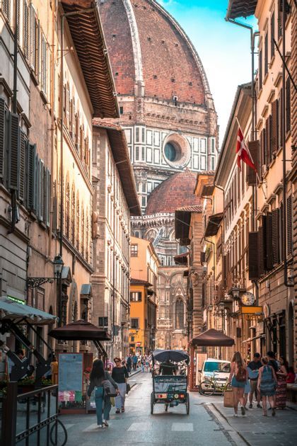 Une vue le long d'une rue étroite et animée bordée de vieux bâtiments, menant vers un grand dôme de cathédrale sous un ciel bleu.