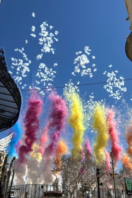 Daylight fireworks create puffs of white smoke and plumes of colorful smoke against a clear blue sky over a city.