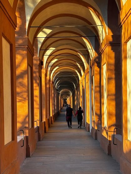 Zwei Personen spazieren entlang eines langen Portikus mit sich wiederholenden Bögen, beleuchtet von goldenem Sonnenlicht, das lange Schatten wirft.