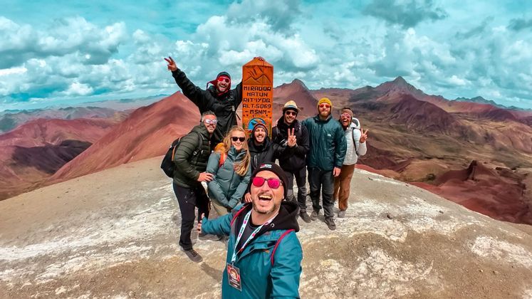 A WeRoad group trip takes a group selfie on a mountain summit with a panorama of colorful red mountains in the background.