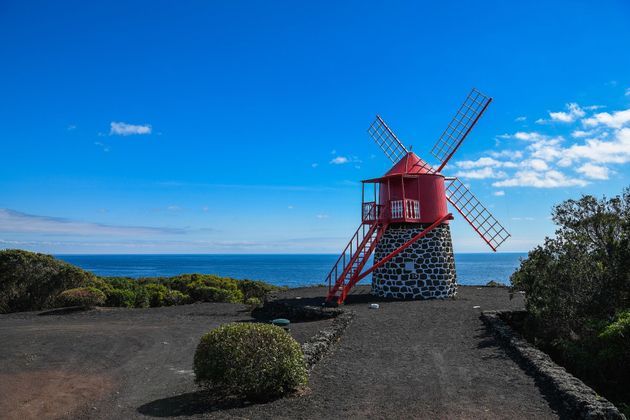 Un mulino a vento rosso con una base in pietra si erge su una costa di terra scura che si affaccia sull'oceano sotto un cielo azzurro brillante.