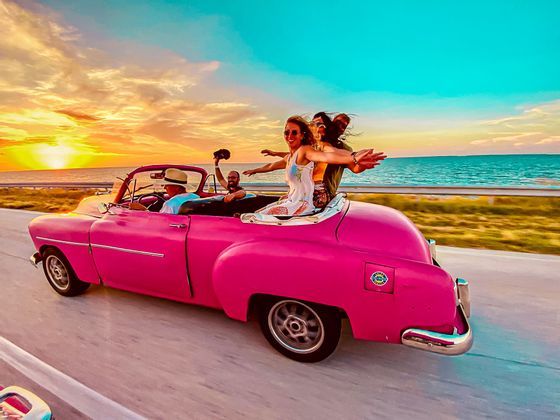 Un viaggio di gruppo WeRoad a bordo di una classica auto decappottabile rosa lungo una strada costiera al tramonto, con le braccia distese.