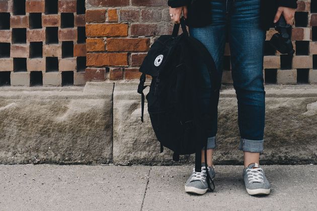 Una persona in jeans arrotolati e scarpe da ginnastica tiene uno zaino nero mentre è in piedi su un marciapiede accanto a un muro di mattoni.