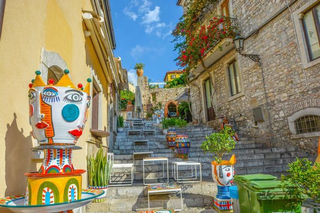 Sculture di teste in ceramica colorate e tavoli da caffè all'aperto disposti su un'ampia scalinata in pietra in un vicolo soleggiato.