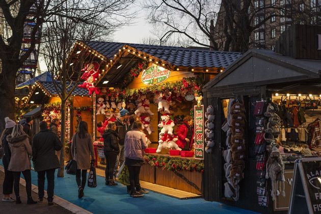 Le persone curiosano tra le bancarelle festive di legno che vendono peluche e regali in un mercatino di Natale all'aperto al tramonto.