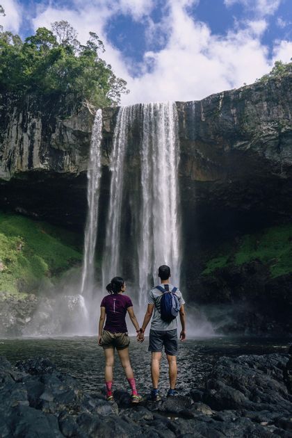Una coppia in abiti da trekking si tiene per mano su rocce bagnate, ammirando un'ampia cascata che scende da una scogliera.