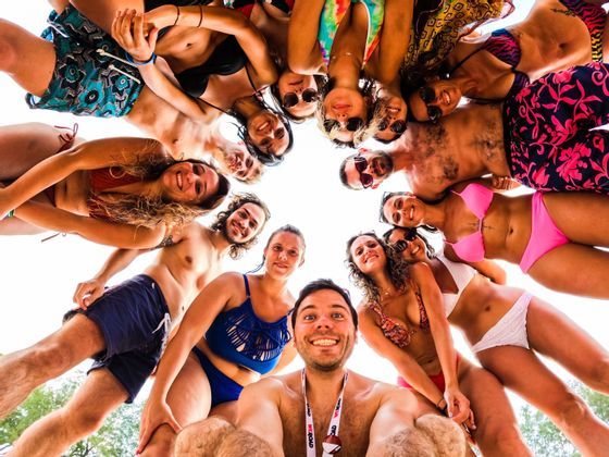 Un gruppo WeRoad in viaggio, in costume da bagno, si raggruppa in cerchio, sorridendo per un selfie dal basso contro un cielo luminoso.