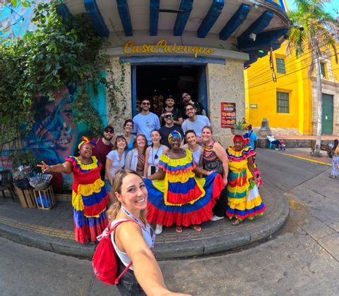 Un viaggio di gruppo WeRoad posa per un selfie con due donne in abiti tradizionali colorati in una strada cittadina.