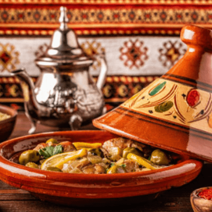 Un tajine traditionnel en terre cuite avec son couvercle entrouvert, révélant un ragoût de viande, d'olives et de citron, avec une théière en argent en arrière-plan.