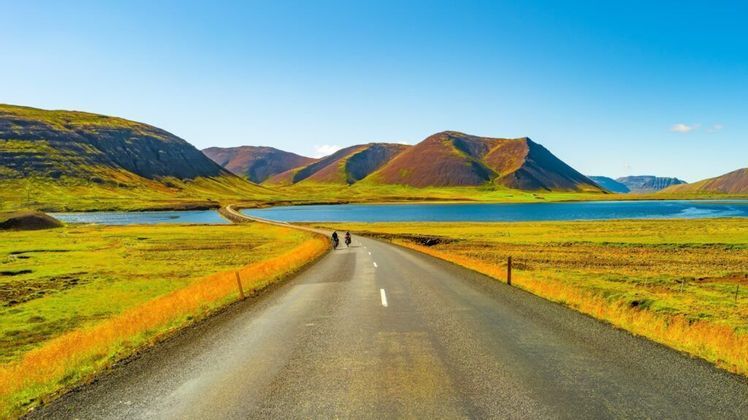 Dos personas en motocicleta recorren una carretera vacía que atraviesa un lago azul en un valle de colinas ondulantes verdes y marrones.