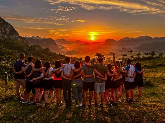 Un groupe WeRoad, bras dessus bras dessous, contemple depuis une colline un coucher de soleil éclatant sur une vallée montagneuse embrumée.