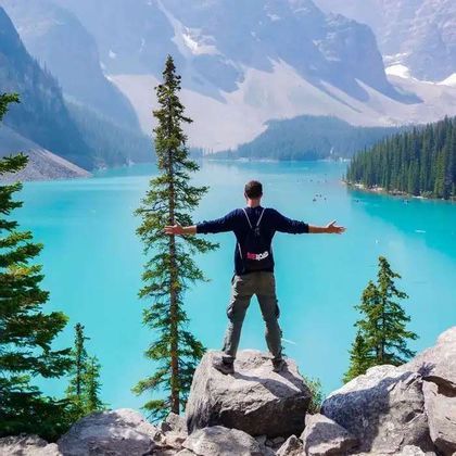 A man with a WeRoad backpack stands on a rock with arms outstretched, viewing a turquoise lake and mountains.