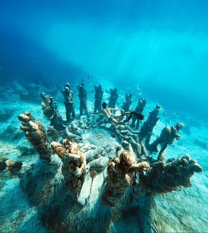Una persona con aletas nada sobre una instalación circular de estatuas submarinas en el arenoso fondo del océano, rodeada de agua azul.