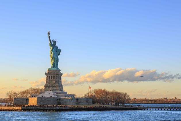 La Statua della Libertà si erge sul suo piedistallo sull'Isola della Libertà, illuminata dalla luce del tramonto contro un cielo azzurro con nuvole.
