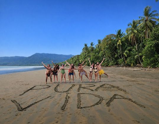 Un grupo de WeRoad posa en una playa tropical detrás de las palabras 'PURA VIDA' escritas en la arena.