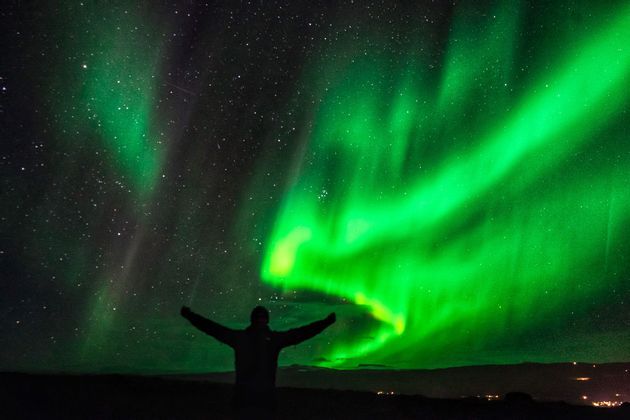 Eine Silhouette einer Person mit ausgestreckten Armen blickt zum grünen Nordlicht in einem sternenklaren Nachthimmel auf.