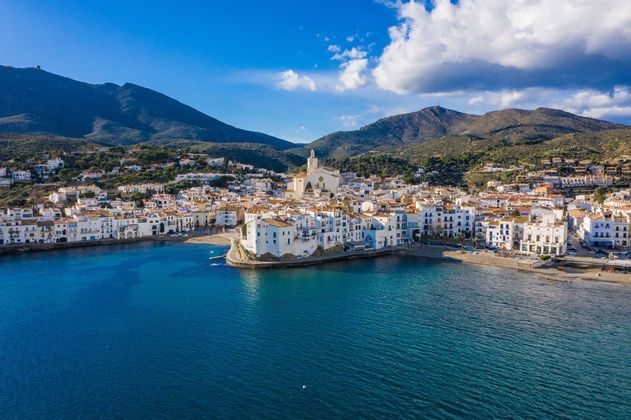 A coastal town with white buildings and a central church is nestled in a bay with blue water and green hills.