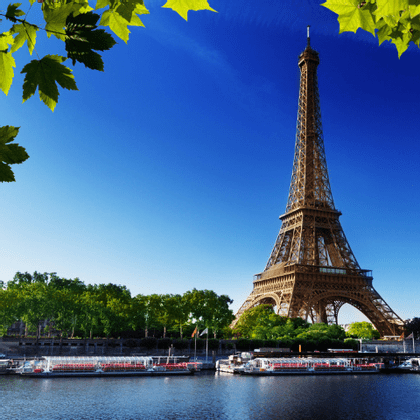 La Torre Eiffel vista da un fiume con barche turistiche ormeggiate, incorniciata da foglie verdi contro un cielo blu.